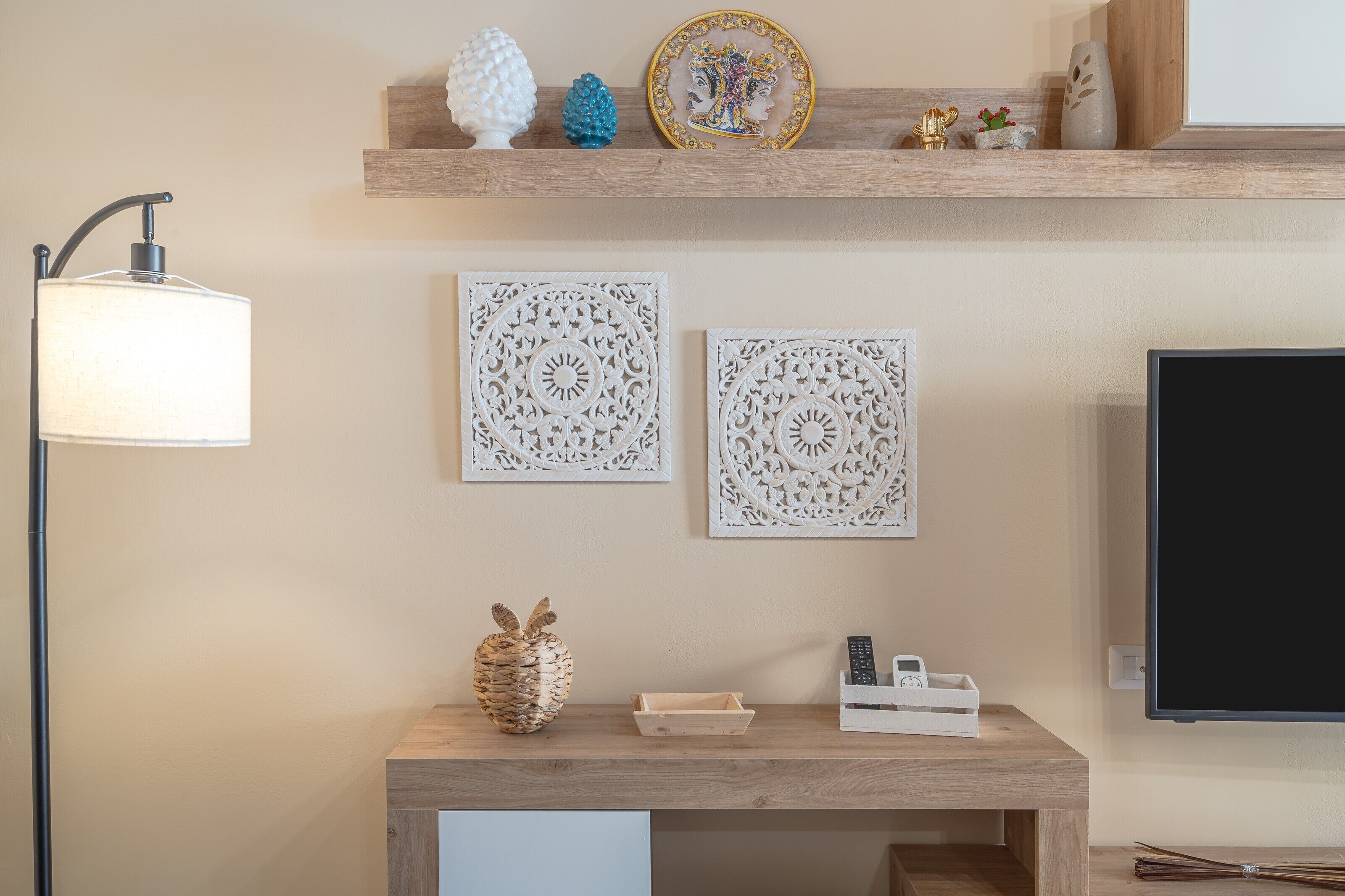 A room with a wooden console table, a wall-mounted TV, decorative wall art, a lamp, and a shelf with various items.