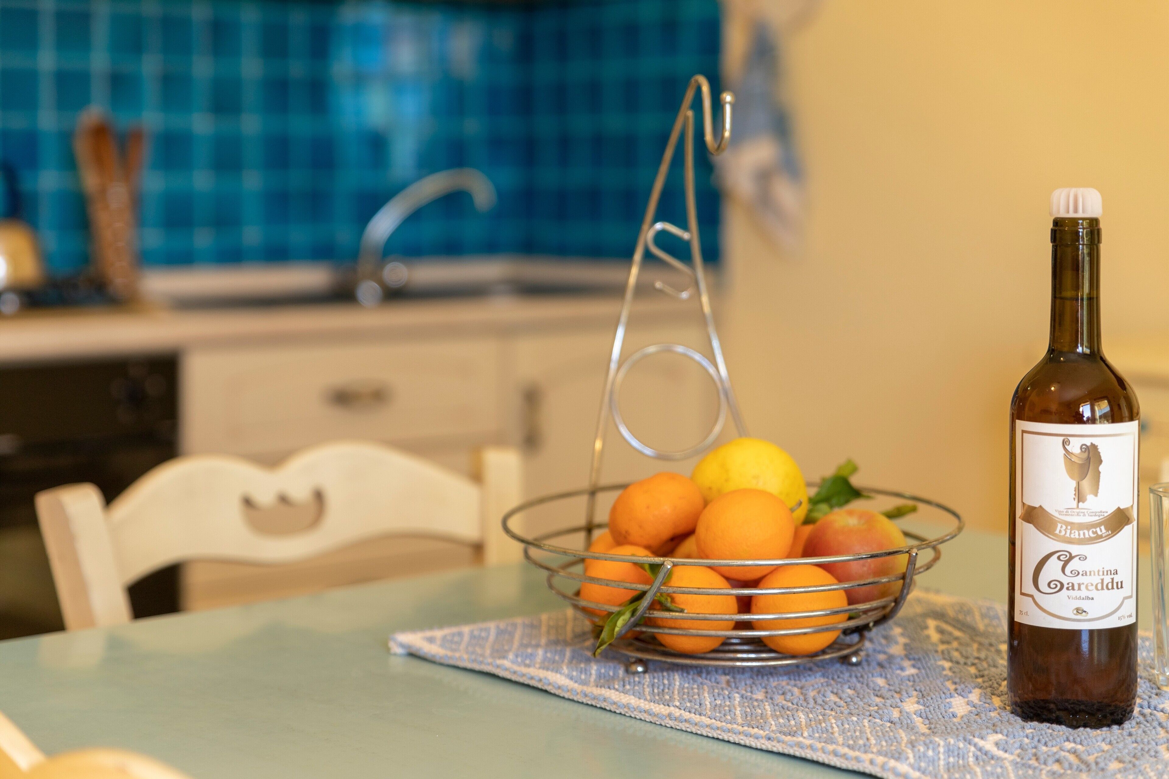 A bottle of wine, a metal fruit basket with oranges and a lemon, and a kitchen setting with a blue tiled backsplash.