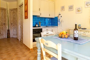 Een keuken met witte kasten, een blauwe betegelde spatwand, een eettafel met stoelen, een fles wijn en een schaal fruit.