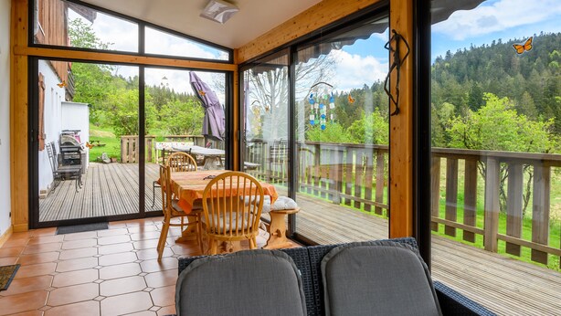 A room with a large sliding glass door leading to an outdoor deck, a wooden table with chairs, and a view of a forested area. - Holiday Home 'Gîte La Ramée' with Mountain View, Private Terrace and Wi-Fi (BAN SUR MEURTHE CLEFCY)