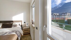 A hotel room with a bed, bedside table, and a view of mountains through a window. - Apartment 'Appartamento Da Jenny' with Mountain View, Balcony and Wi-Fi (aosta)