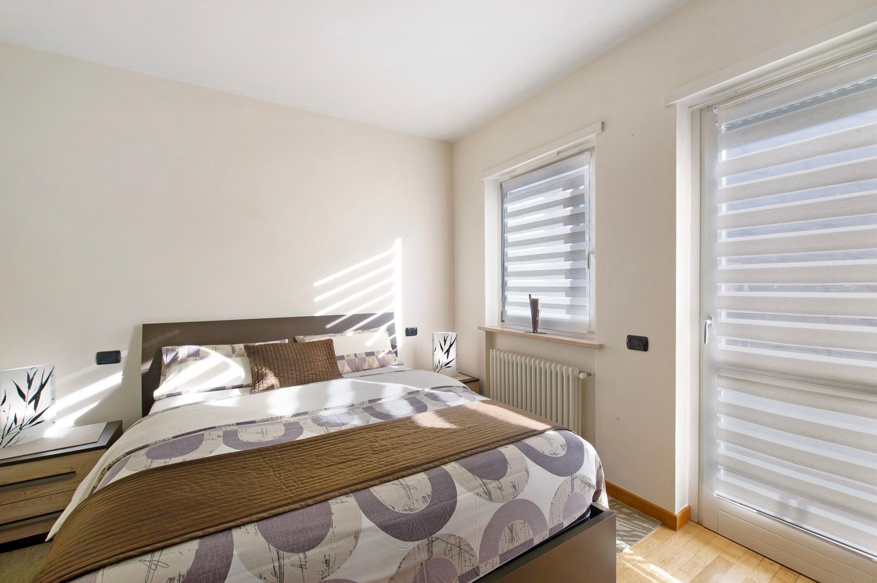 Ein Schlafzimmer mit Bett, Heizkörper, Fenster mit Jalousien und Tür mit Schiebesystem.