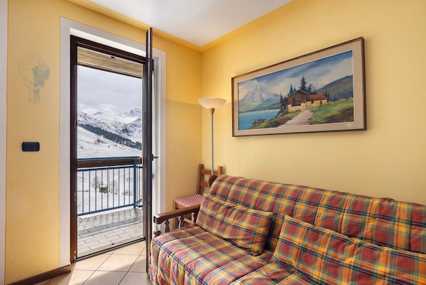 Une pièce avec un canapé à carreaux, un tableau représentant un paysage de montagne et une porte donnant sur un balcon d’où l’on peut admirer des montagnes enneigées.