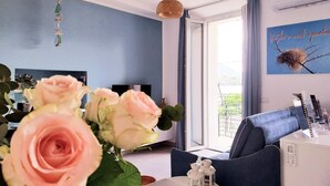 A modern living room with a blue wall, a large window, a sofa, a TV, and a decorative wall piece. - Apartment 'Vichi's Nest' with Lake View, Wi-Fi and Air Conditioning (Pianello Del Lario)