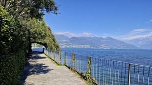 A paved pathway beside a body of water with a metal railing, leading to a distant mountain range. - Apartment 'Vichi's Nest' with Lake View, Wi-Fi and Air Conditioning (Pianello Del Lario)