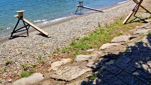 A lakeside view with a pebble beach, two wooden picnic tables, and a clear view of mountains in the background. - Apartment 'Vichi's Nest' with Lake View, Wi-Fi and Air Conditioning (Pianello Del Lario)
