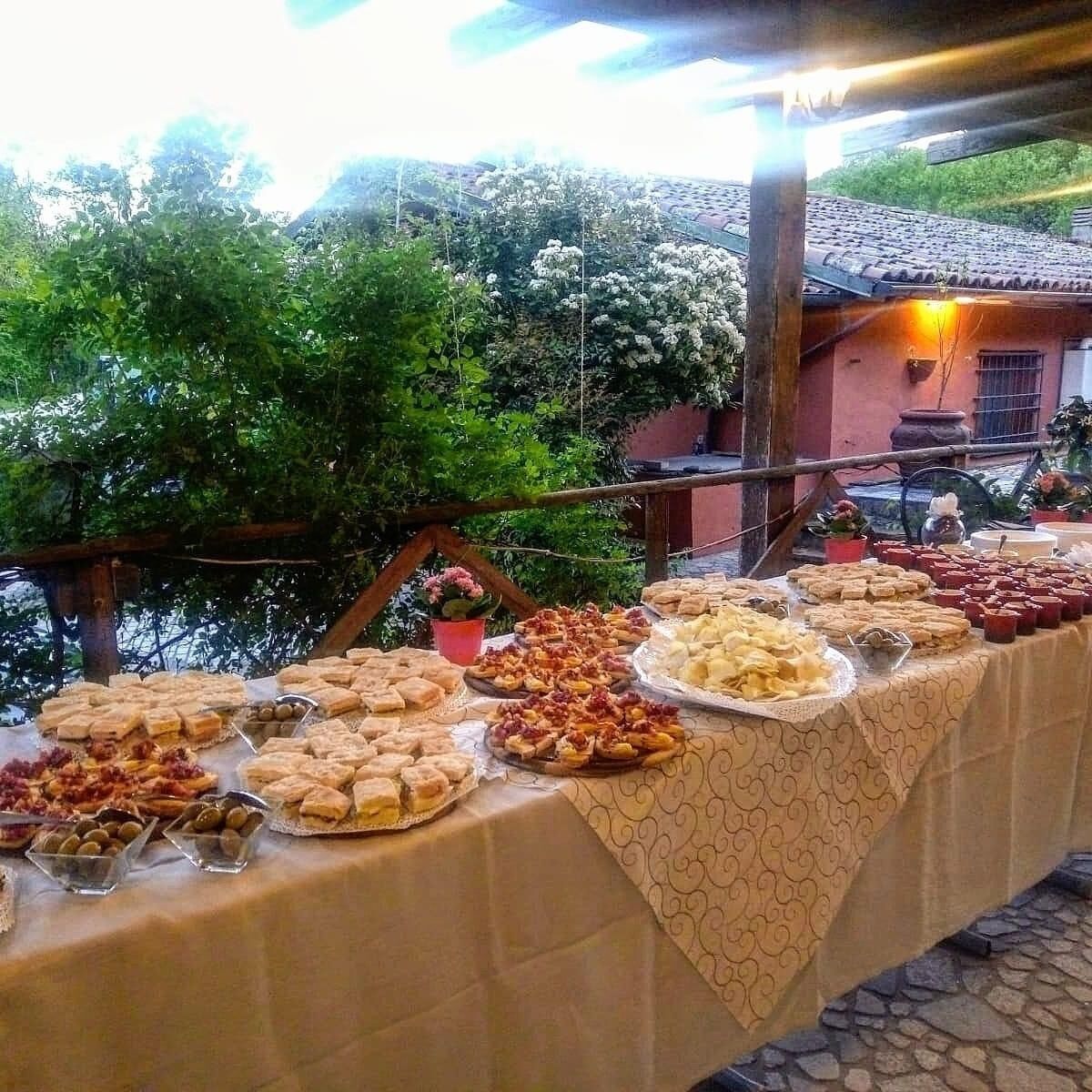 A buffet table with various food items, set outdoors with a view of a red building and greenery in the background.