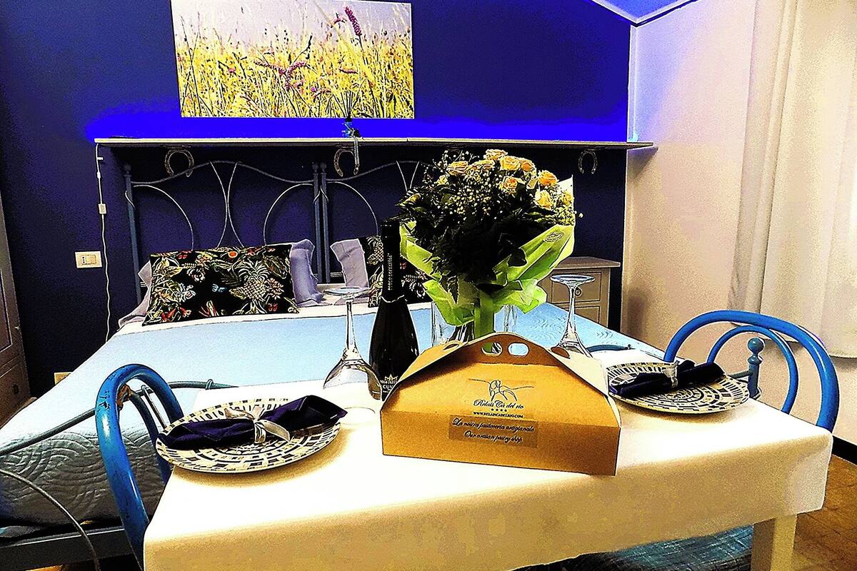 A dining table with a white tablecloth, two blue chairs, a vase with flowers, a bottle, and a box with text.