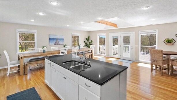 A spacious kitchen with a large island, white cabinetry, and a black countertop. There is a dining area with a table and chairs, and large windows providing natural light. - Huge Lakehouse w/Pool, Hot Tub & Fire Pit Near Nashville! (Hendersonville)