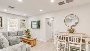 A modern living room with a grey sofa, wooden coffee table, and a dining area with a wooden table and white chairs. - Heated Pool + Backyard: North Palm Beach Retreat! (North Palm Beach)