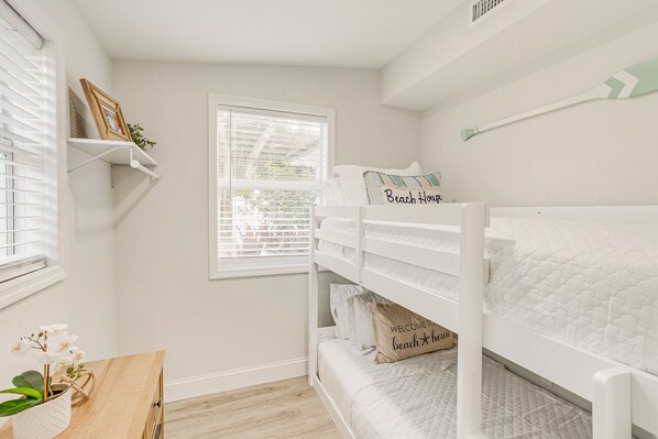 A child's bunk bed with a quilted mattress, a small wooden table with a plant, and a window with blinds.