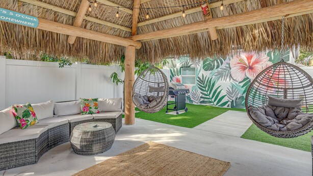 A covered outdoor seating area with a thatched roof, wicker furniture, and hanging chairs. - Heated Pool + Backyard: North Palm Beach Retreat! (North Palm Beach)