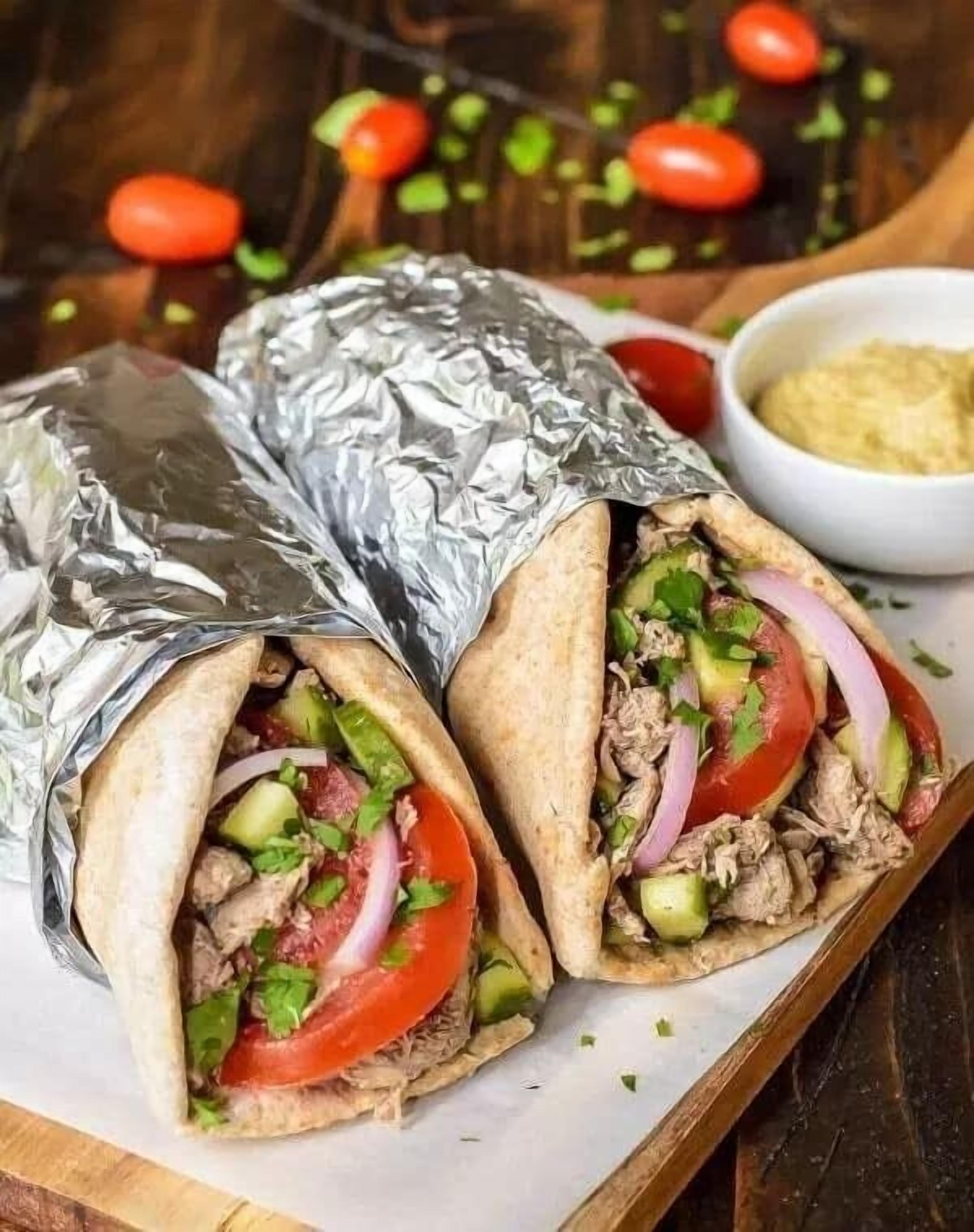 A wrap sandwich with meat, avocado, tomato, and onion, served on a wooden board with foil.