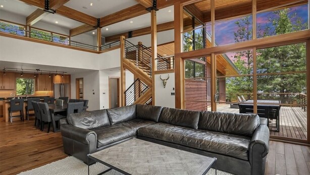 A spacious living area with a leather sectional sofa, wooden beams, a staircase, and large windows offering a view of the outdoors. - Rinky Dink Retreat - Suncadia LUXE With MIL Suite and Hot Tub (Cle Elum)