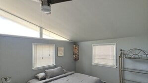 A bedroom with a bed, a nightstand, a desk, a chair, a ceiling fan, and a window with blinds. - Peace in the City (Decatur)