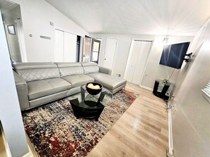 Un salon avec un canapé gris, une table basse noire sur laquelle se trouve un bol, un tapis à motifs et un téléviseur posé sur un meuble.