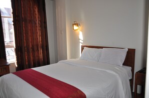 A neatly made bed with white linens and a red blanket, a wall-mounted light fixture, and a window with curtains.