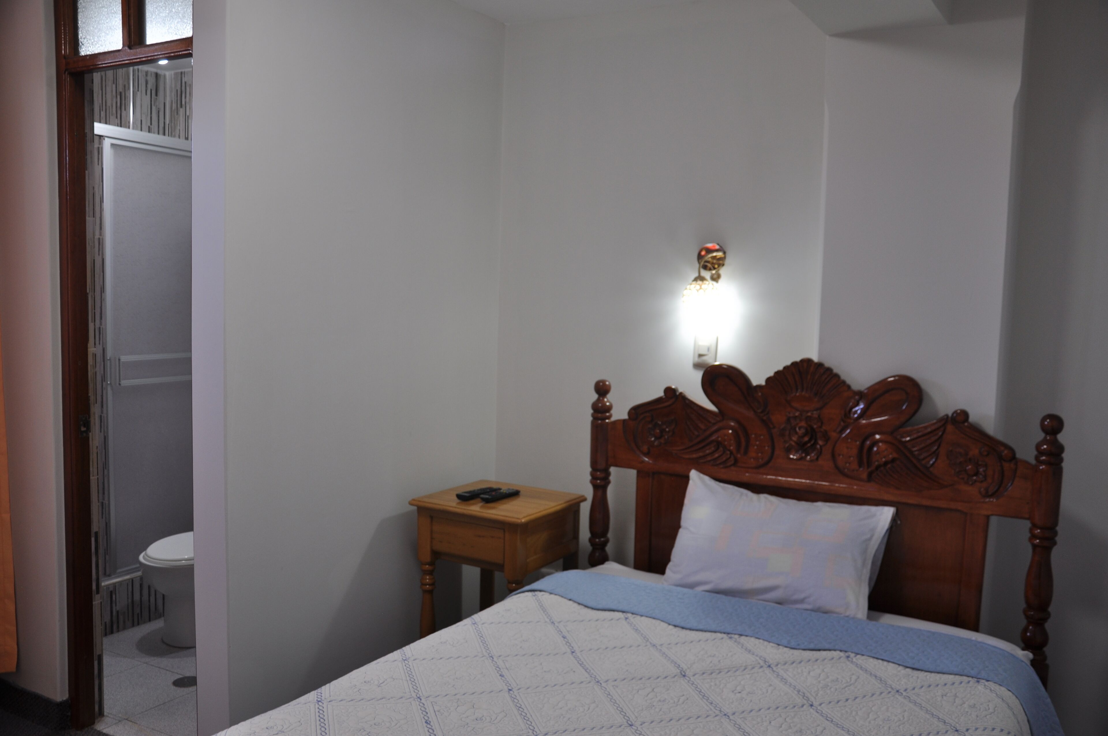 A bedroom with a wooden bed, a bedside table, a nightstand, a wall-mounted light, and a view of a bathroom.