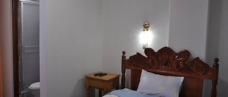 A bedroom with a wooden bed, a bedside table, a nightstand, a wall-mounted light, and a view of a bathroom.