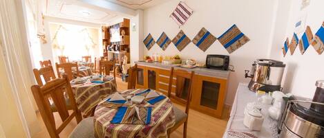 Uma área de refeições com mesas e cadeiras de madeira, cozinha com micro-ondas, cafeteira e objetos decorativos na parede.