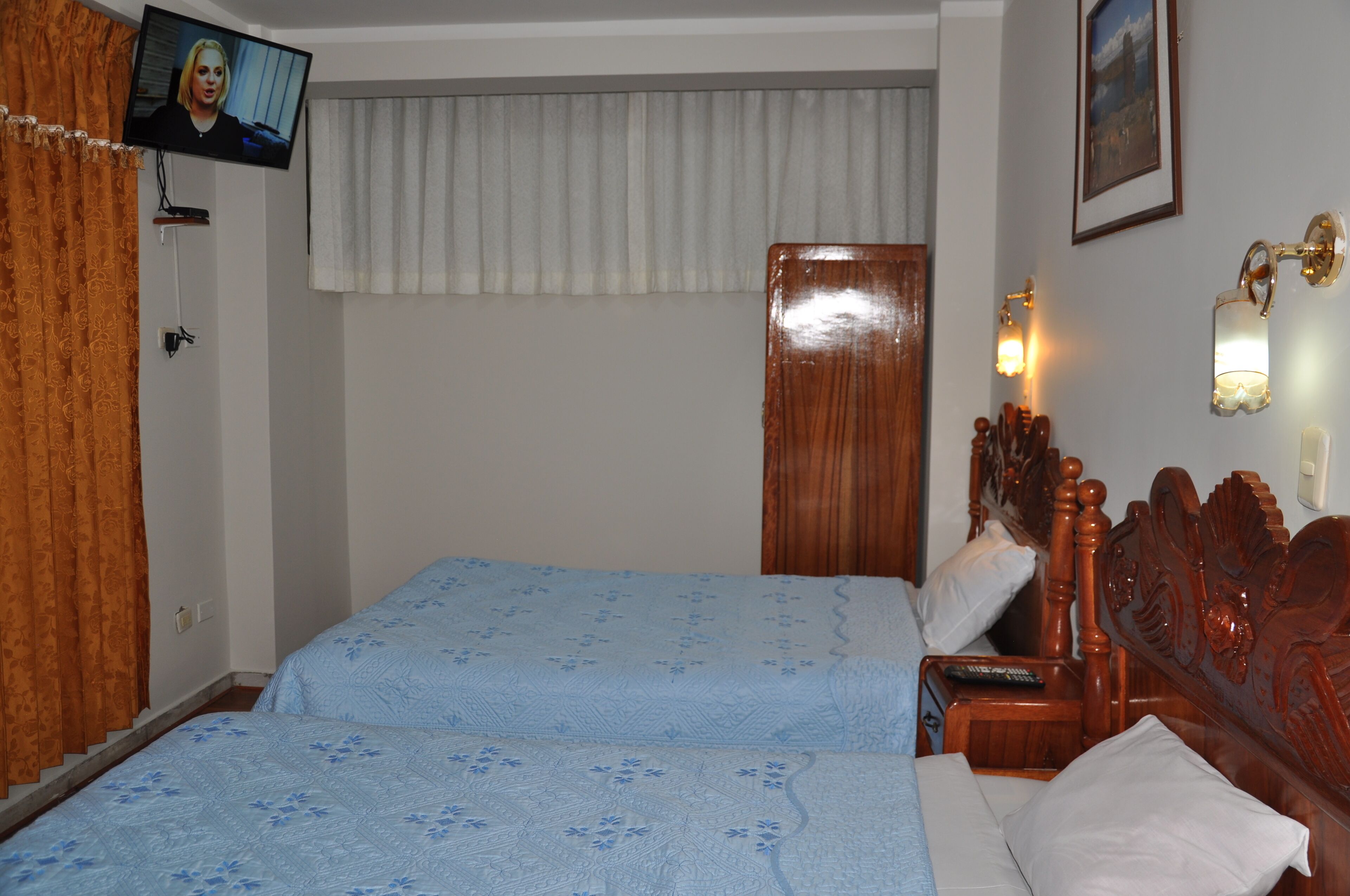 Una habitación de hotel con dos camas, un televisor montado en la pared, un cabecero de madera y una mesita de noche con una lámpara.