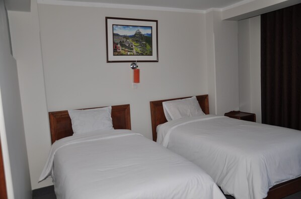 A hotel room with two single beds, a wooden headboard, a framed picture on the wall, and a window with curtains.
