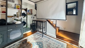 Un interior de hogar con una pantalla de proyector, una escalera de madera, un tapete estampado y un mueble de madera gris con objetos decorativos.
