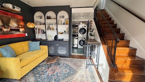 Una sala de estar acogedora con un sofá amarillo, un tapete estampado y una escalera que lleva a un área de lavandería.