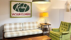 A living room with a checkered sofa, a green armchair, a wooden coffee table, a lamp, and a sign that reads "Kirby Vacuum Cleaners".