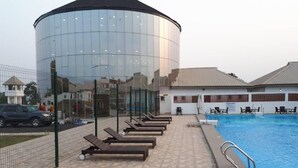 Zona de piscina con sillones de descanso, un edificio de cristal y un estacionamiento cercado.