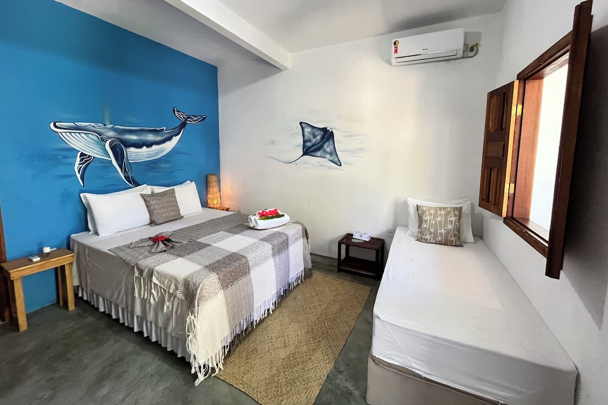 A bedroom with a bed, a bedside table, a nightstand, a wall-mounted air conditioner, and a window with wooden shutters.