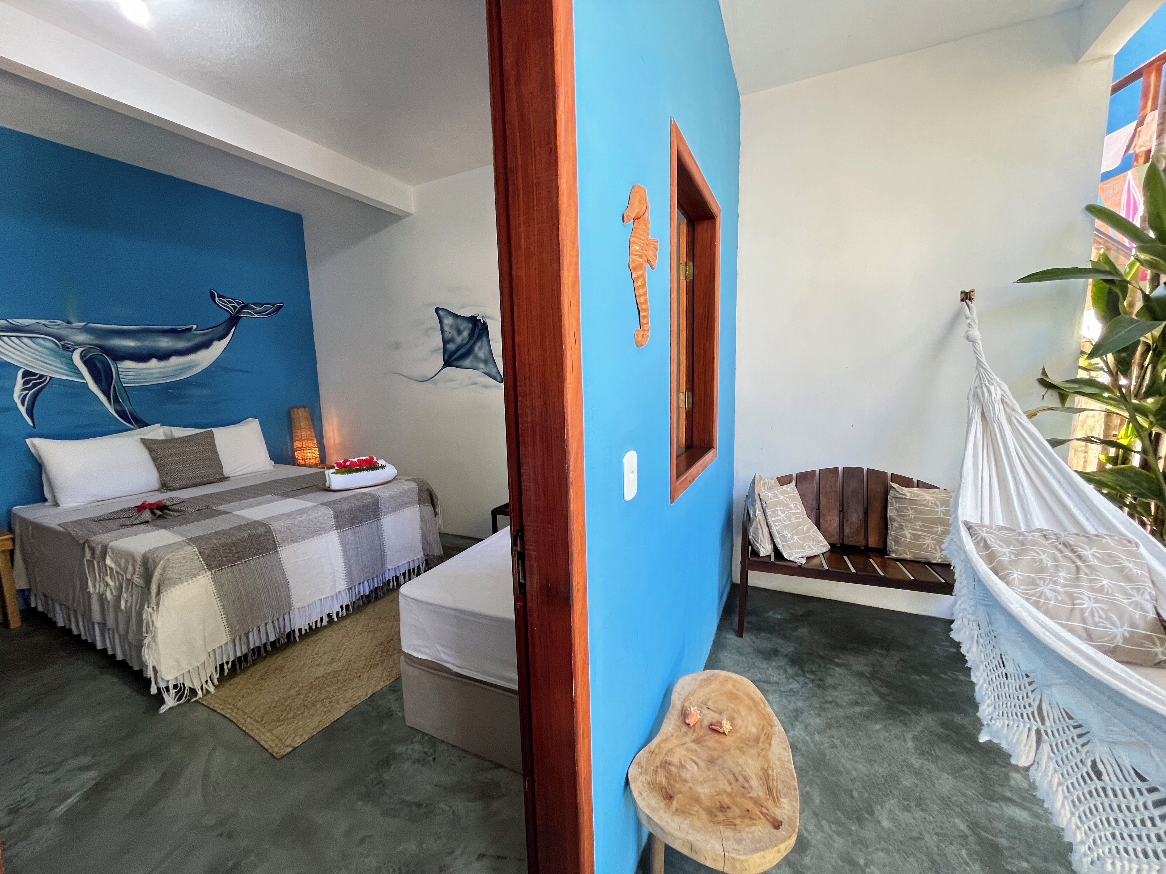 Un dormitorio con cama, una hamaca, un taburete de madera y un mural en la pared.