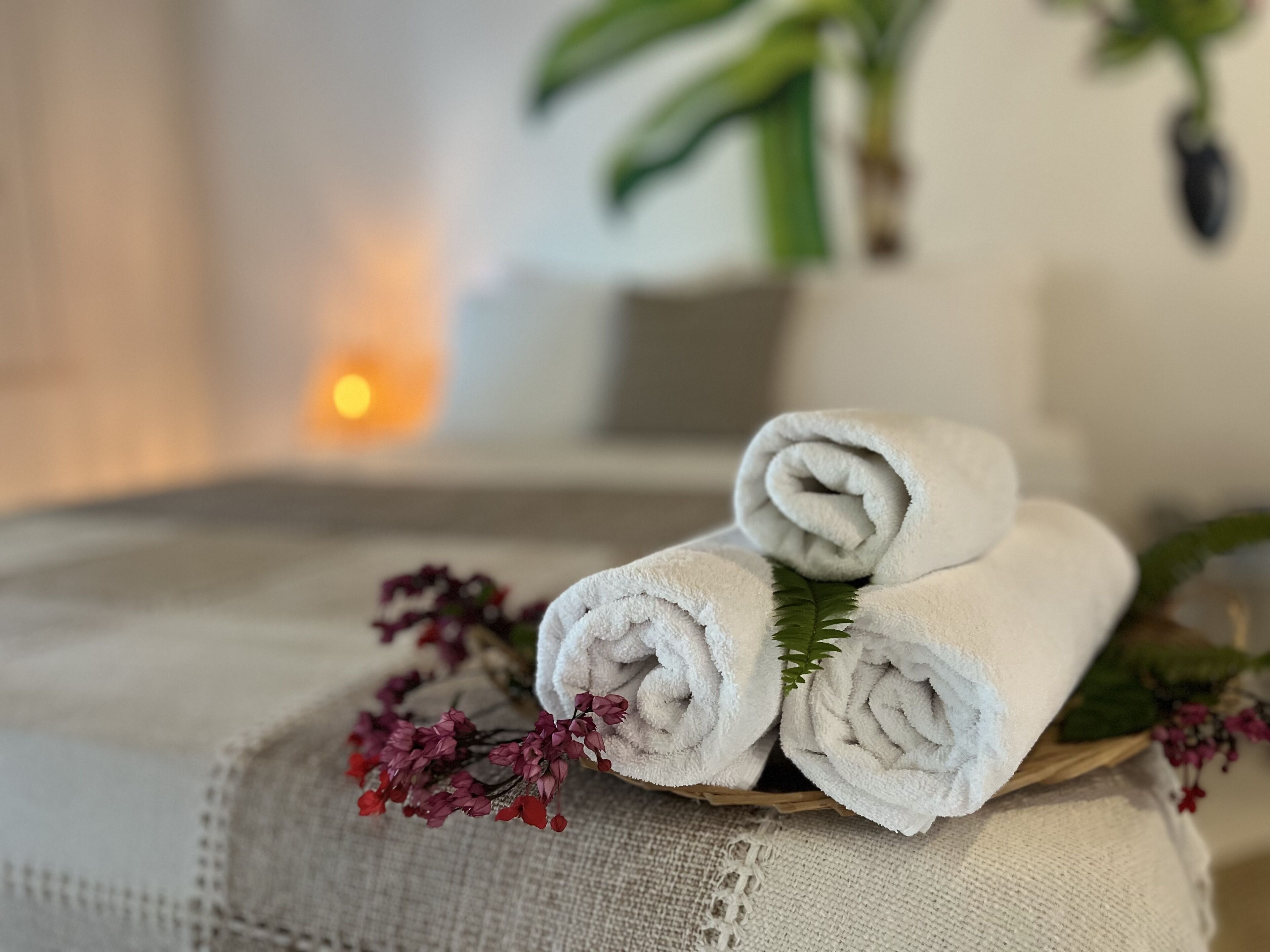 Een netjes opgestapelde stap witte handdoeken op een oppervlak van gestructureerde stof, samen met kleine rode bloemen en een groen blad, met een wazige achtergrond die een knus interieur doet vermoeden.