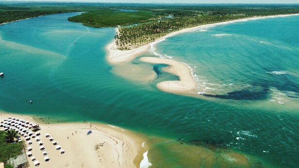 An aerial view of a coastal area with a sandy beach, clear blue waters, and a small island with dense vegetation. - Pousada Nascente do Sol (Cairu)