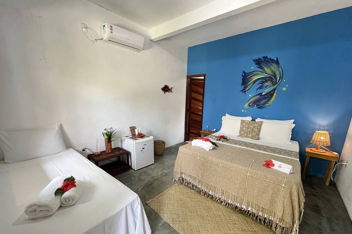 A hotel room with a bed, a smaller bed, a wall-mounted air conditioner, a small table with a plant, a nightstand, and a wall painting.