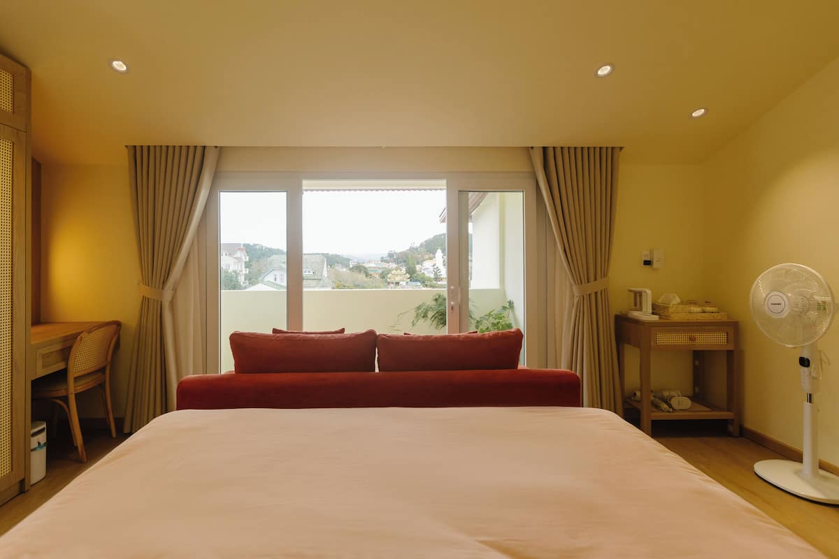 A hotel room with a large bed, a desk, a chair, a bedside table with a lamp, a fan, and a window with curtains.