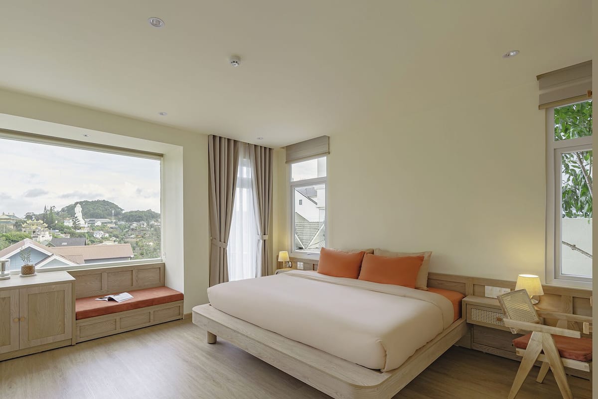 A bedroom with a large window, a bed, a bedside table, a nightstand, a bench, and a potted plant.