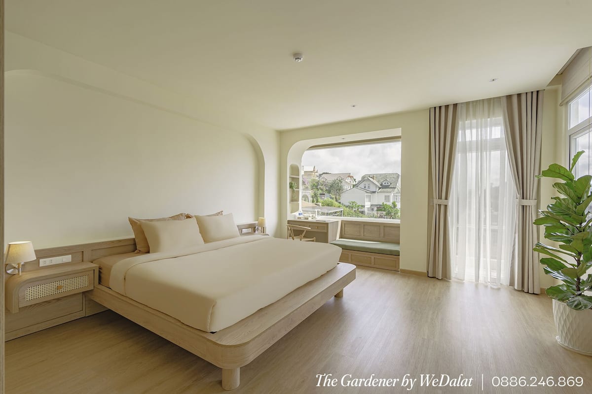 A spacious bedroom with a large bed, a bedside table, a window with curtains, and a view of buildings outside.
