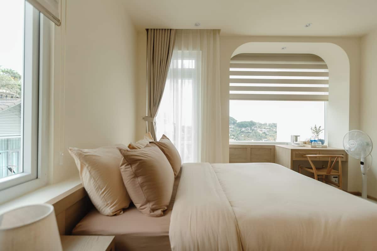 A bedroom with a large bed, a desk, and a chair. There is a window with curtains, a lamp, and a fan.