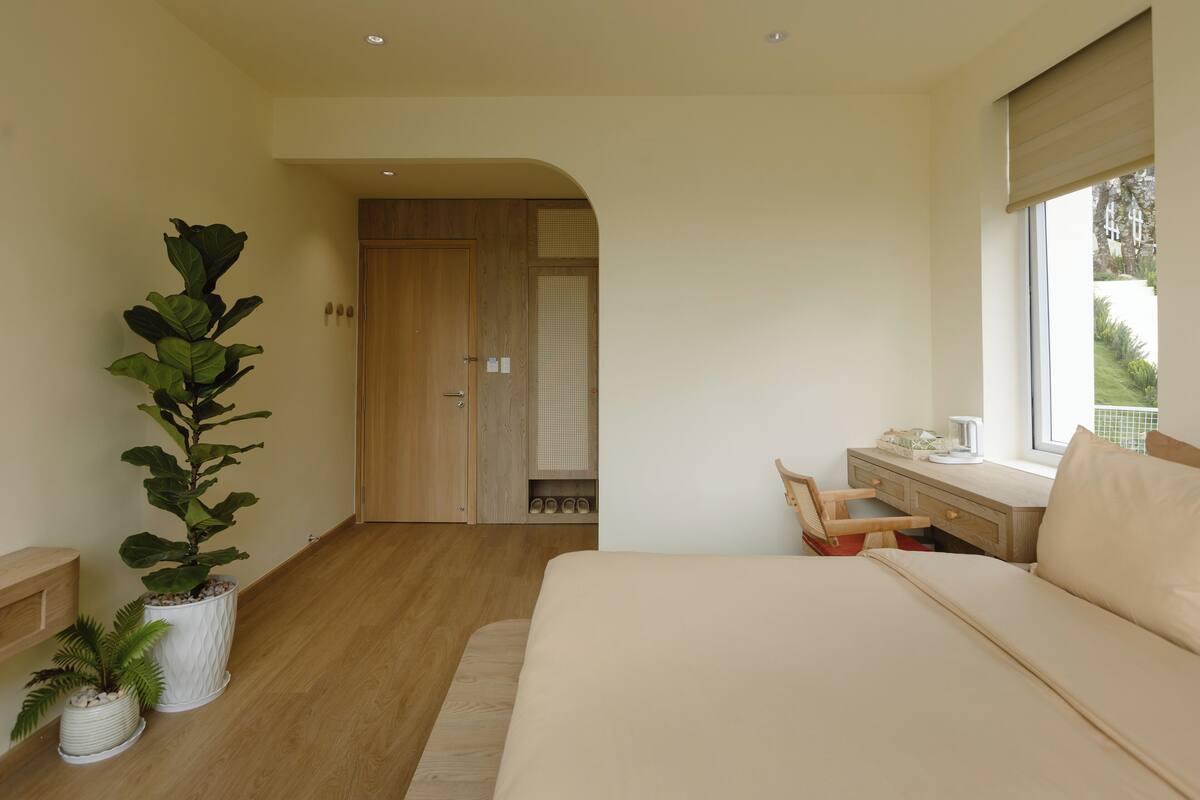 A bedroom with a bed, a desk, a window, and a potted plant.
