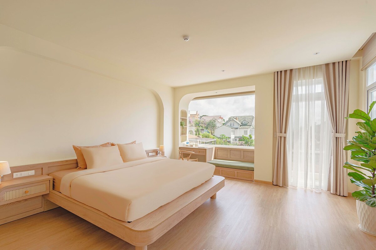 Ein geräumiges Schlafzimmer mit einem großen Bett, einem Nachttisch, einem Fenster mit Vorhängen und Blick auf Gebäude.