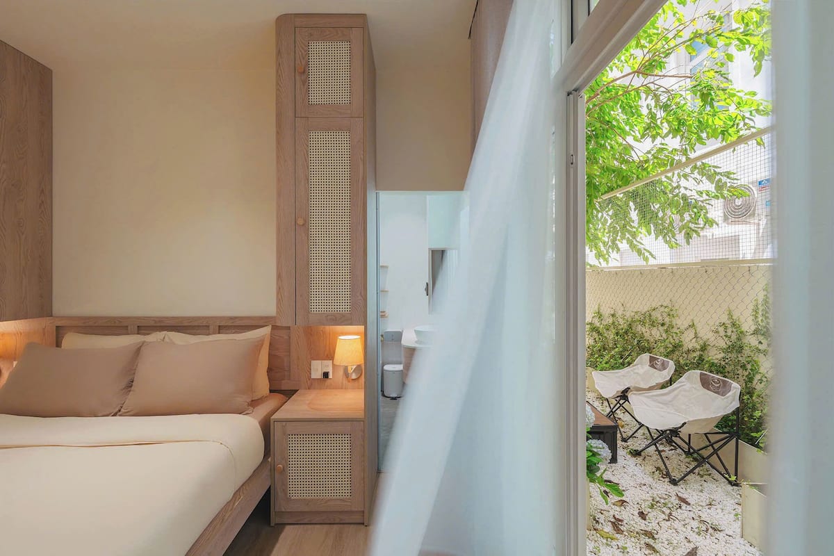 A bedroom with a wooden headboard, a bedside table with a lamp, and a view of an outdoor seating area with chairs and plants.
