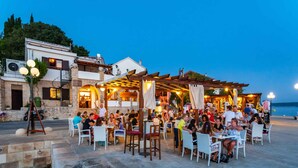 Un área de comedor al aire libre con piso de piedra y sillas blancas, rodeada de edificios con fachadas de piedra y pérgolas de madera.