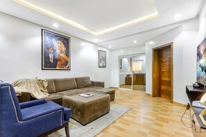 Una sala moderna con un sofá modular, un sillón azul y una mesa de centro. Hay cuadros en las paredes y una puerta de madera que da a la cocina.
