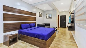 Een moderne slaapkamer met een houten hoofdbord, een groot bed met blauw beddengoed, een klein bureau, een stoel en een aan de muur bevestigde televisie.