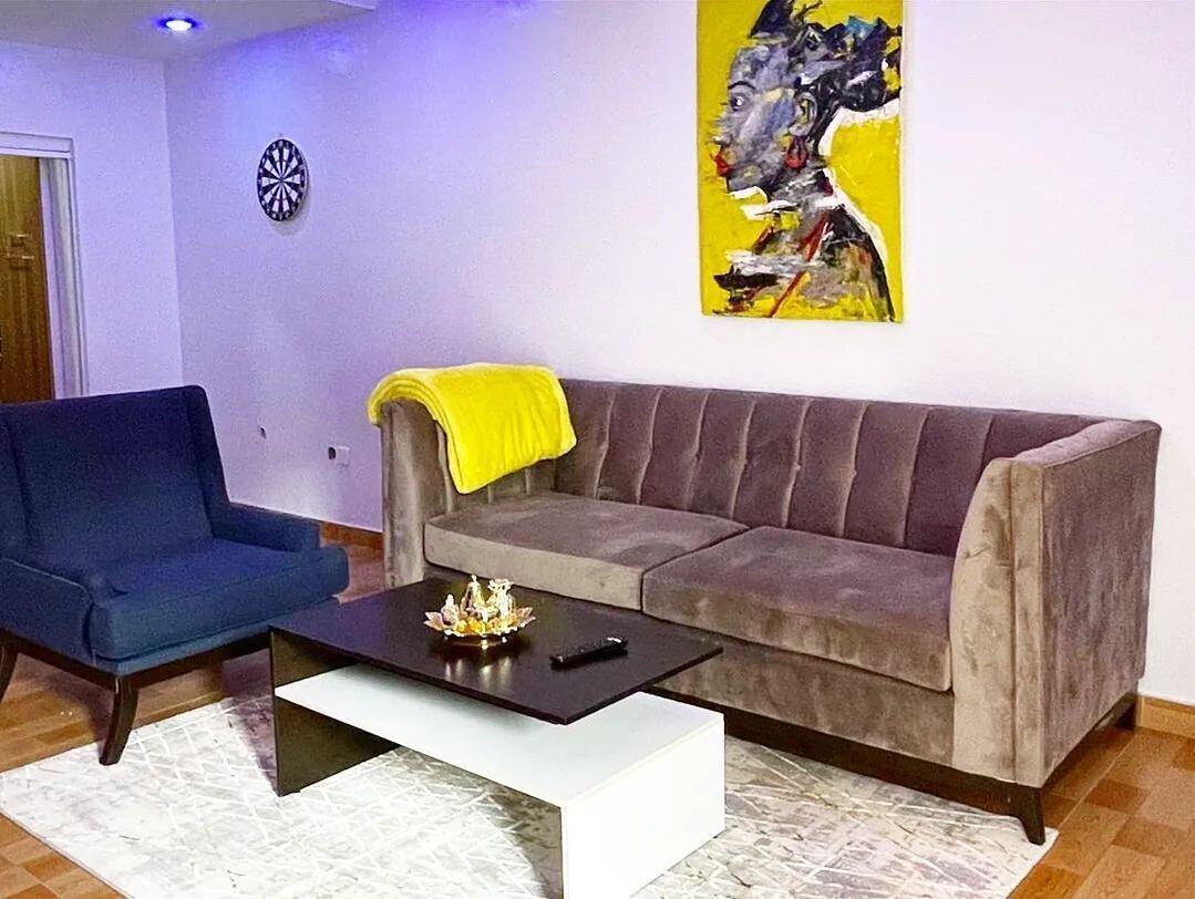 A living room with a blue armchair, a grey sofa with a yellow throw, and a coffee table with a remote control.