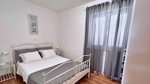 Een enkel bed met metalen frame, witte beddengoed en een hoofdbord. Er staat een nachtkastje met een lamp en een kleine tafel met een lamp. De kamer heeft houten vloeren en een raam met grijze gordijnen.