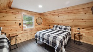 Un dormitorio con paredes de madera, una cama con colcha a cuadros, una mesita de noche, un espejo redondo y una ventana con vista a la vegetación.
