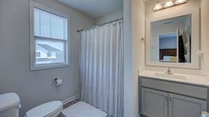 Een badkamer met een toilet, douchegordijn, wastafel en spiegel, en een raam.
