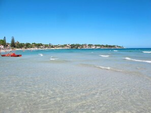A beach with clear shallow waters, a red boat, and a distant shoreline with buildings.